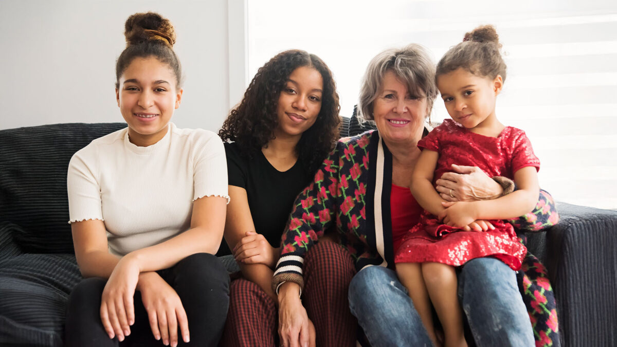 Groupe familial multigénérationnel assis sur un sofa, trois filles et une femme plus âgée posant et souriant ensemble.