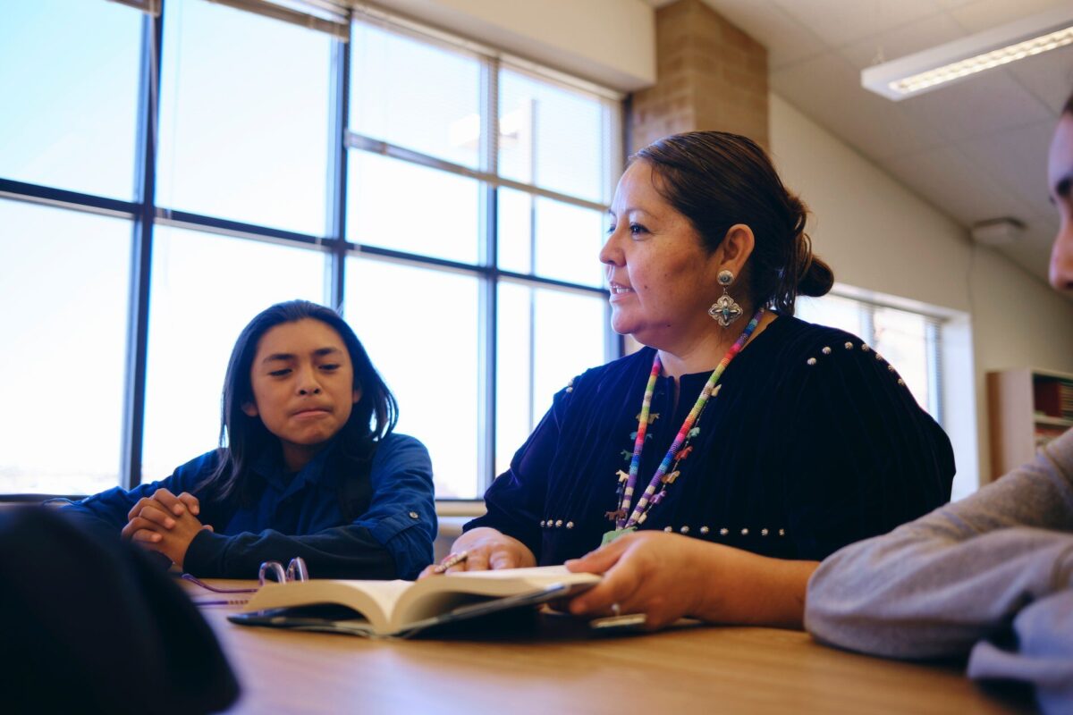 Une enseignante autochtone discute avec un adolescent autour d’une table, un livre ouvert devant eux, dans une salle de classe lumineuse.