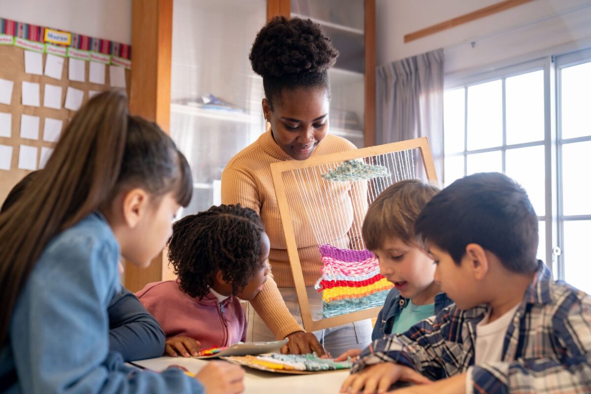 Enseignante montrant un métier à tisser avec une création colorée à un groupe d’enfants attentifs autour d’une table dans une salle de classe.