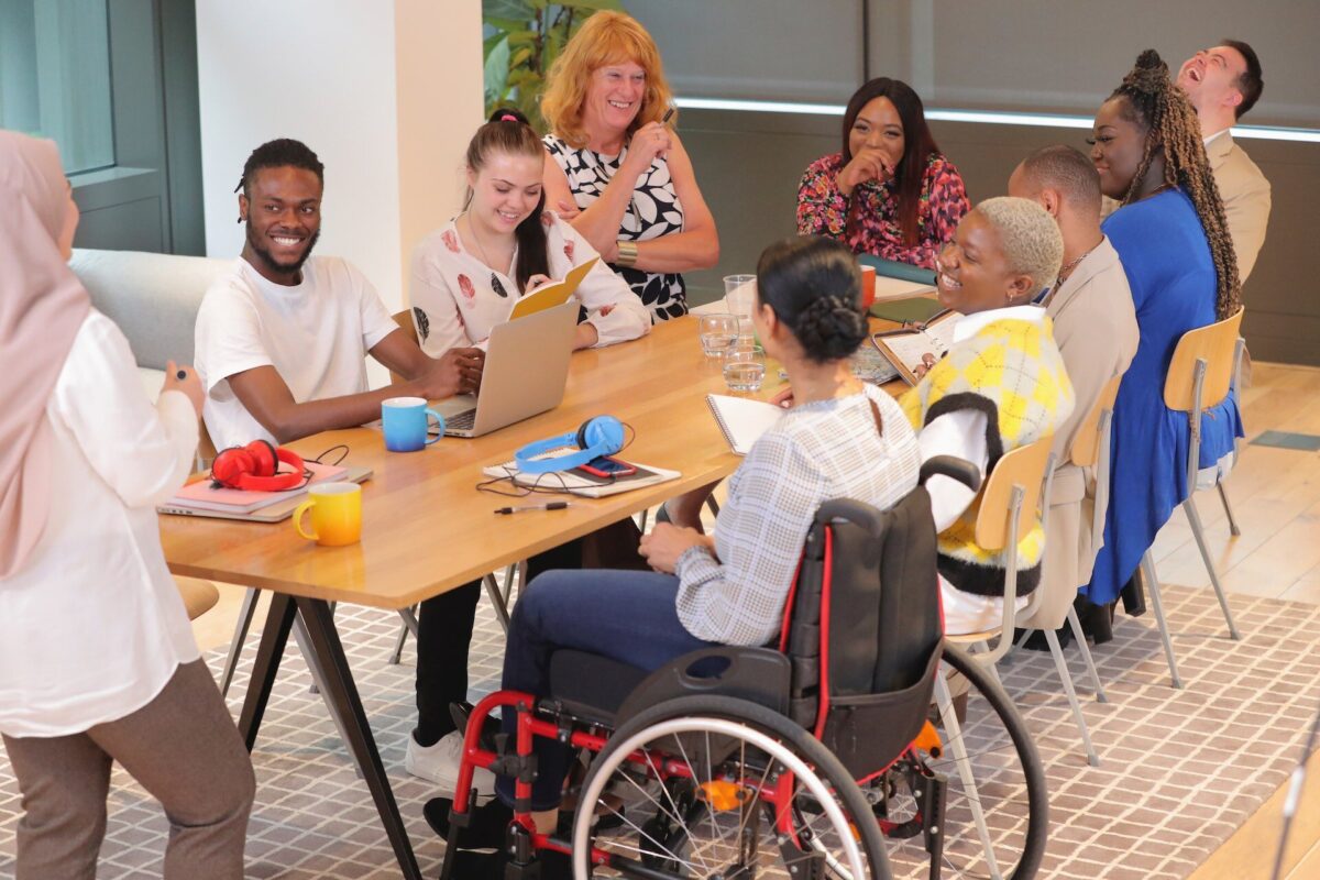 Groupe de collègues diversifiés assis autour d’une grande table en réunion informelle, souriants et en pleine discussion, incluant une personne en fauteuil roulant.