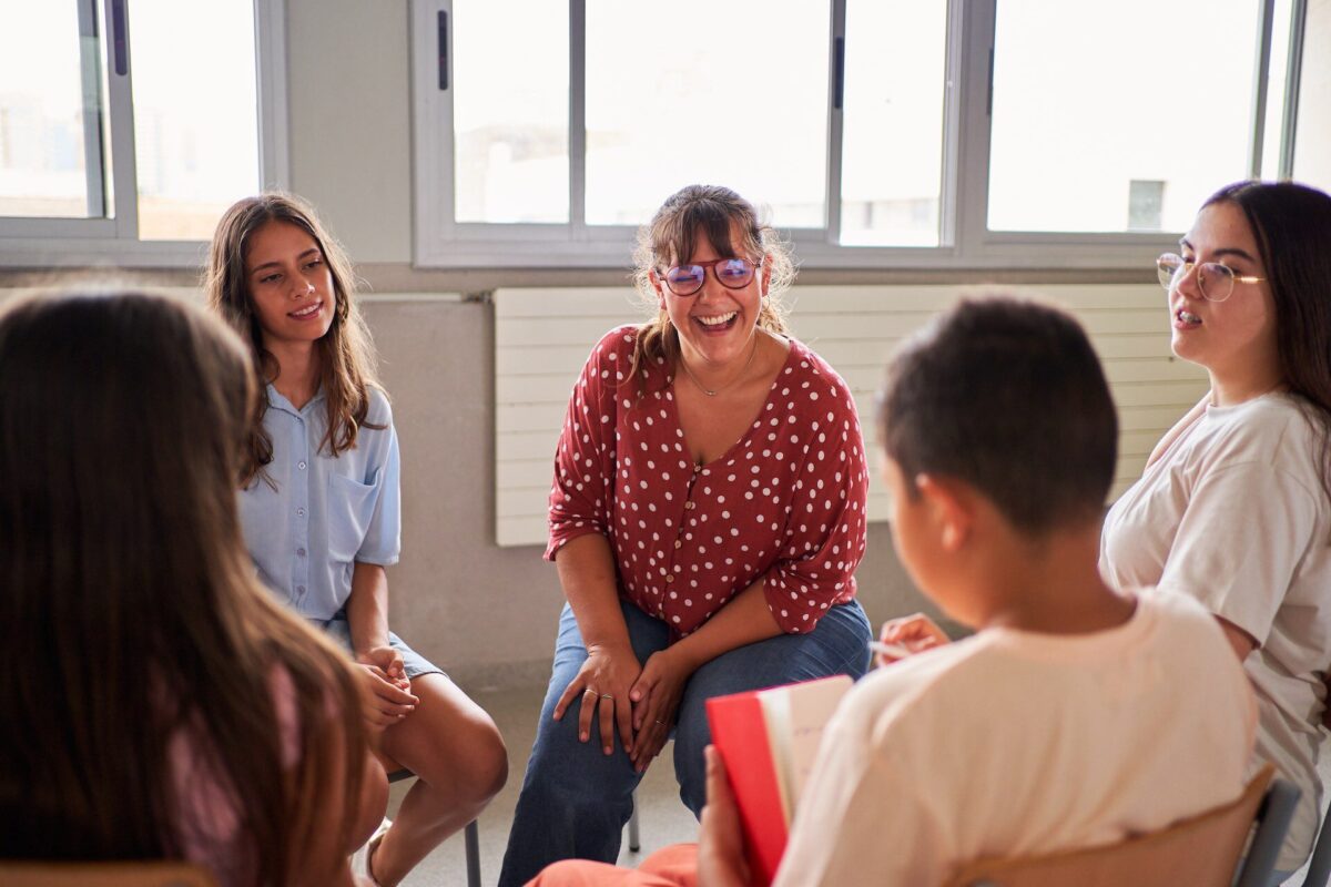 Groupe d’adolescents et une adulte assis en cercle dans une salle lumineuse, discutant ensemble; l’adulte au centre rit avec le groupe dans une ambiance détendue.